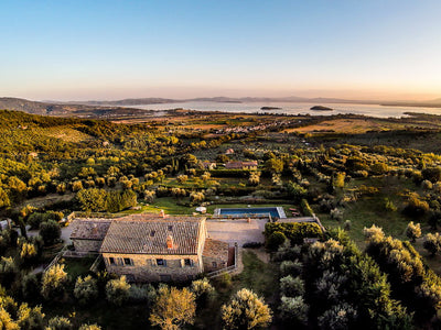 Aerial view of a Cortona villa surrounded by greenery with a pool, set against a scenic landscape.