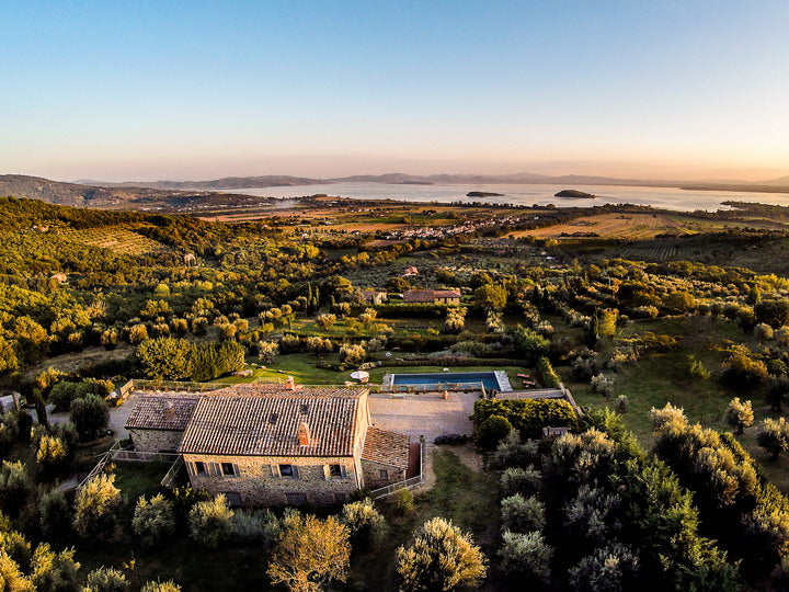 Aerial view of a Cortona villa surrounded by greenery with a pool, set against a scenic landscape.